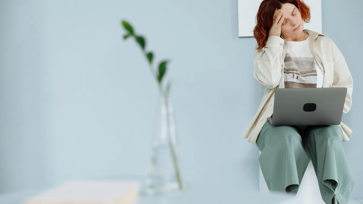 Woman sitting with laptop, holding head in a minimalist space, depicting stress and fatigue.