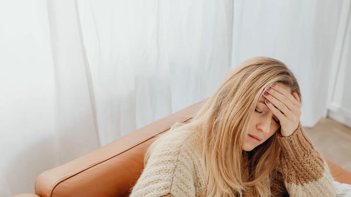 A woman in a knitted sweater sits with a headache on a cozy sofa indoors.