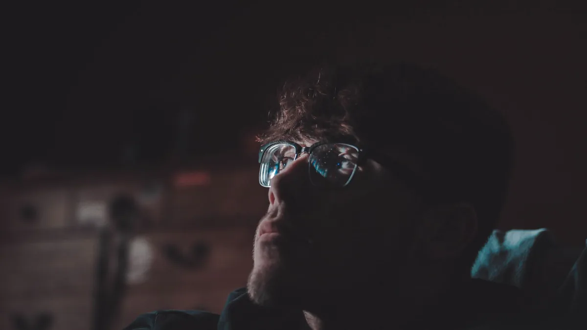 Dark and reflective portrait of a man wearing eyeglasses in a dimly lit room, capturing thoughtful expression.