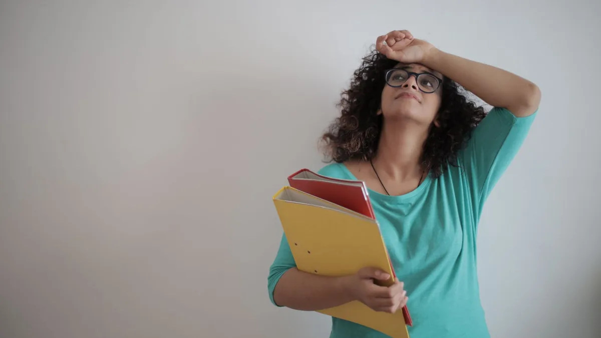 Low angle of unhappy exhausted woman in glasses and casual clothes with folders of papers looking up with sorrow and touching forehead while.