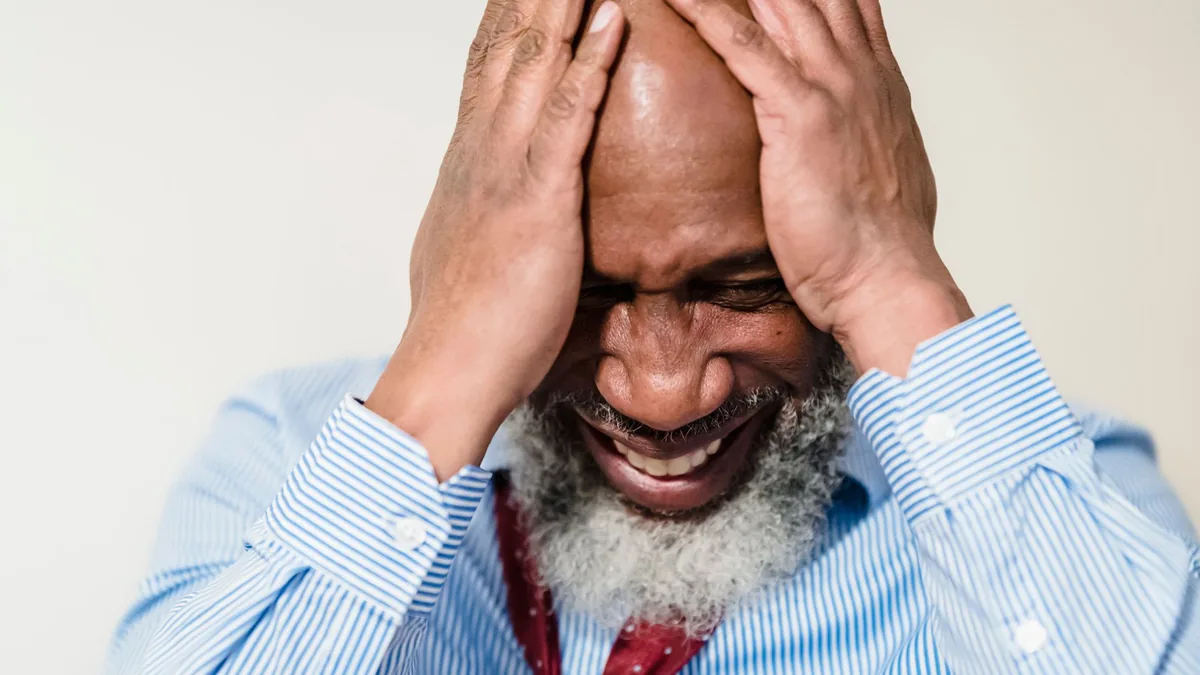 Elderly man in striped shirt showing stress and emotion with hands on head.