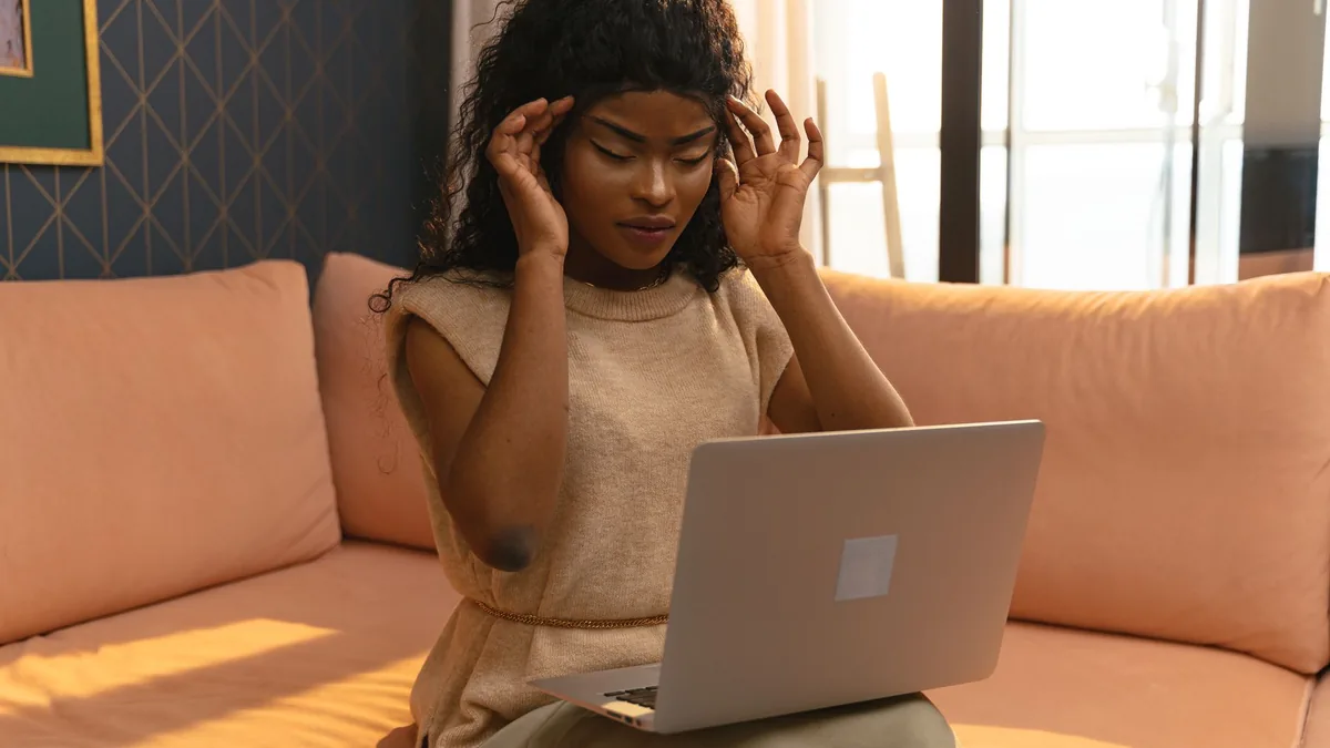 A young woman holding her head in pain while working on a laptop in a cozy home interior.