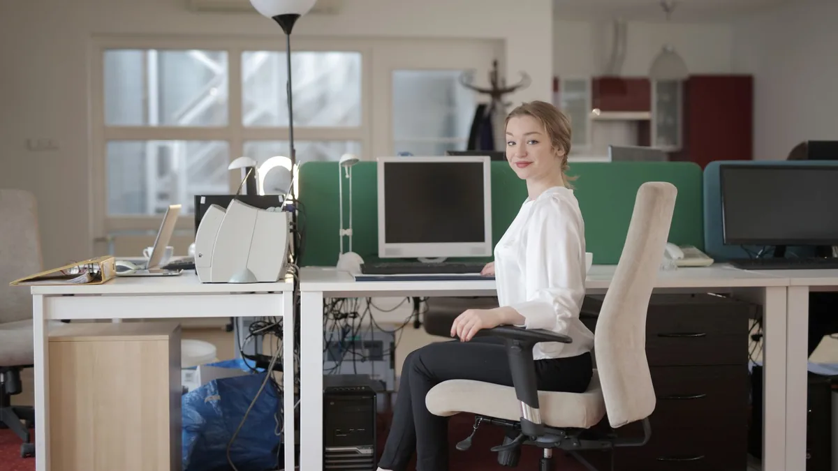 Side view of glad female worker in classy outfit sitting at table on office chair during work in modern workplace and looking at camera.