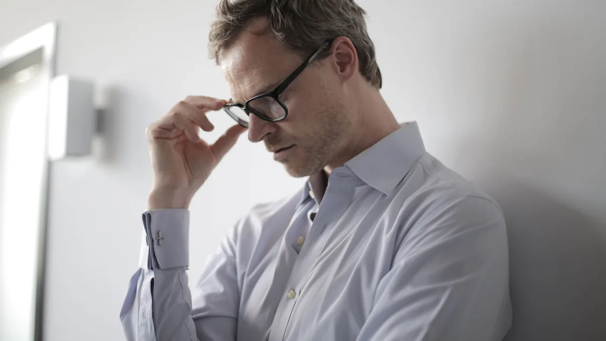 An adult man in a shirt and glasses appears stressed while leaning against a wall indoors.