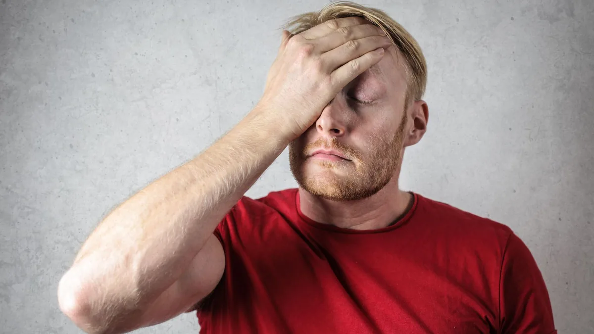 A frustrated man in a red shirt holds his head in stress against a neutral background.